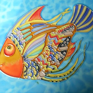 May include: A colorful ceramic tile featuring a stylized fish with orange, yellow, blue, green, and red details. The fish has a large, round eye and intricate patterns on its body and fins.