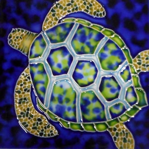 May include: A ceramic tile featuring a sea turtle design. The turtle's shell is a mosaic of blue and green hues, outlined in light blue. The flippers and head are a speckled pattern of tan and olive green, set against a deep blue background.