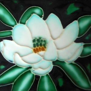 May include: A square ceramic tile featuring a raised relief design of a white magnolia flower with light blue shading on the petals. The center of the flower is green and yellow. The background consists of dark green leaves against a black field.