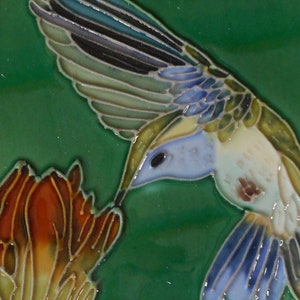 May include: A square ceramic tile featuring a detailed hummingbird design. The bird has blue, white, and green feathers, a long beak, and is feeding from a red and orange flower against a green background.