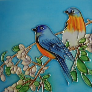 May include: A square ceramic tile featuring two colorful birds perched on a flowering branch against a light blue background. The birds have blue, orange, and white plumage. The flowers are white and the leaves are green.