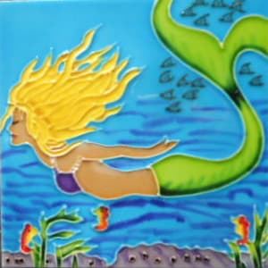 Puede incluir: Un azulejo de cerámica con una ilustración pintada a mano de una sirena con el pelo rubio suelto y una cola verde. La sirena nada en agua azul con un banco de peces pequeños y coral.