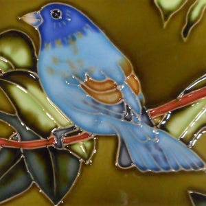 Könnte beinhalten: Keramikfliese mit einer detaillierten Illustration eines blauen Vogels, der auf einem Ast mit grünen Blättern sitzt. Der Vogel hat blaue und braune Federn und einen weißen Schnabel. Der Hintergrund ist eine Mischung aus Grün und Braun.