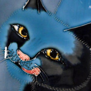 May include: A ceramic tile depicting a cat's face. The cat has a blue head, black features, and yellow eyes. The nose is pink, and the mouth is red. The tile has a glossy finish and a decorative design.