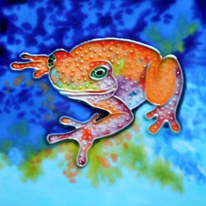 May include: A colorful ceramic tile featuring a bright orange frog with green eyes and white spots. The frog is painted in a vibrant style with pink, blue, and yellow accents. The background is a blue and green watercolor-like design.