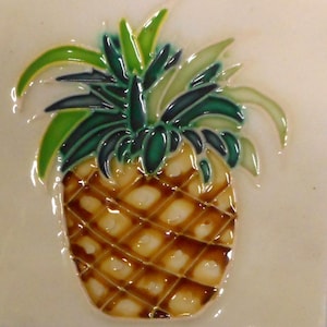 Pode incluir: Azulejo de cerâmica com um desenho de abacaxi. O abacaxi tem um corpo castanho texturizado com um padrão de grade e uma coroa de folhas verdes e amarelas. O azulejo tem um acabamento brilhante.