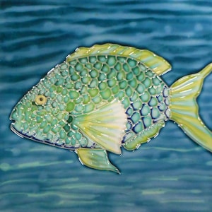 May include: A ceramic tile featuring a colorful fish design. The fish has a green body with a scale pattern, yellow fins, and a white patch. The background is a blue water pattern.