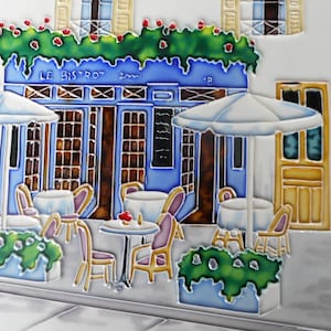 Może przedstawiać: Płytka ceramiczna w kolorze niebieskim i białym przedstawiająca paryski bistro z ustawionymi na zewnątrz stolikami i krzesłami. Płytka zawiera napis "Le Bistro" i zielone winorośl z czerwonymi kwiatami.