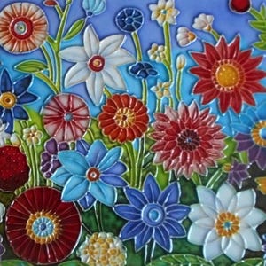 Op de afbeelding: Een decoratieve keramische tegel met een levendig, veelkleurig bloemenpatroon tegen een lichtblauwe achtergrond. Het ontwerp bevat diverse gestileerde bloemen in tinten rood, wit, blauw en geel, met verhoogde contouren die elk bloemblad omlijnen.