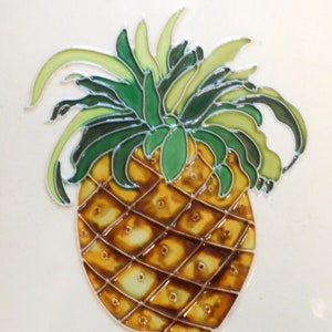 May include: A hand-painted ceramic tile featuring a pineapple with a green leafy top and a yellow and brown fruit. The pineapple is painted in a realistic style with details on the fruit and leaves.