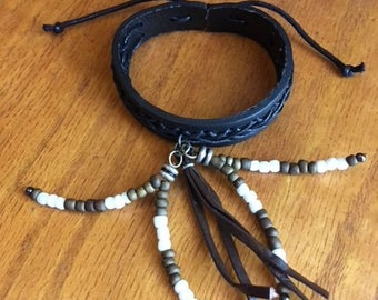 Può includere: Un braccialetto in pelle nera con un design intrecciato e un fiocco in pelle marrone. Il braccialetto è decorato con perline bianche e marroni.