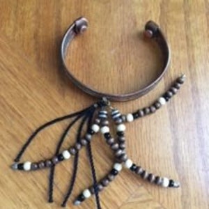 Peut inclure: Un bracelet de manchette en cuivre avec un charme suspendu composé de perles en bois brun, de perles noires et de glands noirs.