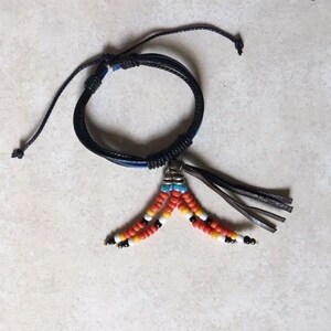 Peut inclure: Un bracelet en cuir noir avec un pendentif en perles. Le pendentif est compos&eacute; de perles orange, blanches et bleues.