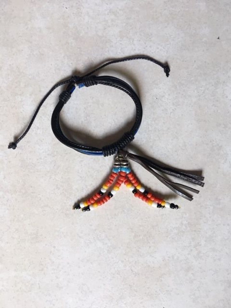 Peut inclure: Un bracelet en cuir noir avec un pendentif en perles. Le pendentif est compos&eacute; de perles orange, blanches et bleues.