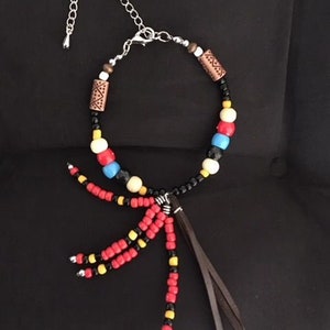 Peut inclure: Un collier de perles avec un cordon noir et un pompon en cuir. Les perles sont rouges, jaunes, bleues, blanches et brunes. Les perles brunes ont un motif géométrique.