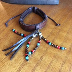 Peut inclure: Un bracelet en cuir marron avec un pompon de perles. Les perles sont brunes, rouges, bleues et blanches.