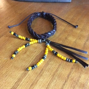 Peut inclure: Un bracelet en cuir marron avec un design tressé et un pompon de perles jaunes et noires.