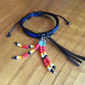 Peut inclure: Un bracelet en cuir noir avec une bande bleue et un pompon perl&eacute;. Le pompon est compos&eacute; de perles rouges, blanches, noires et jaunes.