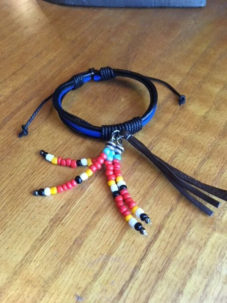 Peut inclure: Un bracelet en cuir noir avec une bande bleue et un pompon perl&eacute;. Le pompon est compos&eacute; de perles rouges, blanches, noires et jaunes.