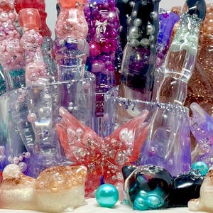 以下が含まれることがあります： 蝶や猫の置物、様々な装飾品など、カラフルな樹脂アートのコレクション。赤、青、紫、透明色で、キラキラやパールのアクセントが施されています。一部のアイテムは透明な容器に入れられています。