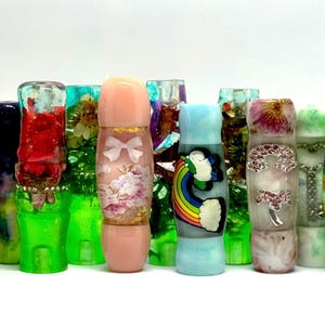 Könnte beinhalten: Eine Sammlung farbenfroher, dekorativer Harz-Flaschenverschlüsse. Zu den Designs gehören Blumenmuster, ein Regenbogen und Glitzerakzente. Die Verschlüsse sind in verschiedenen Farben erhältlich, darunter Rosa, Grün und Blau.