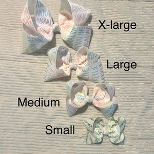 May include: Four white hair bows in varying sizes: X-large, Large, Medium, and Small. The bows have a pastel rainbow design with names printed on them. The bows are arranged on a light wood-grain surface.