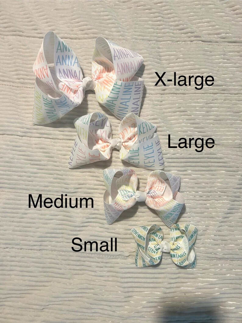 May include: Four white hair bows in varying sizes: X-large, Large, Medium, and Small. The bows have a pastel rainbow design with names printed on them. The bows are arranged on a light wood-grain surface.