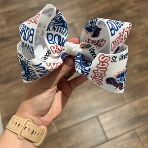 May include: White hair bow with a repeating pattern of the words "Knights" and "St. Vince" in blue and red. The bow features a football helmet graphic. The bow is held in a hand.