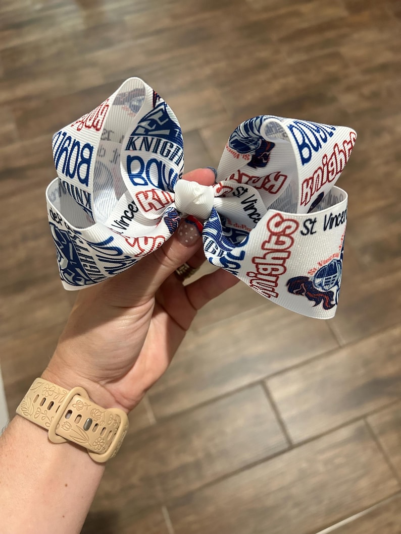 May include: White hair bow with a repeating pattern of the words "Knights" and "St. Vince" in blue and red. The bow features a football helmet graphic. The bow is held in a hand.