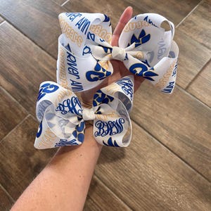 May include: Three white hair bows with blue and gold accents. The bows feature the text "River Ann Cougars" and "BHS" in a repeating pattern. The bows are made of a ribbon material.