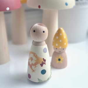 May include: Wooden peg dolls with painted details. One doll has a cream-coloured dress with a chicken design and colourful polka dots. Another doll has a yellow mushroom cap with white dots. Other mushroom-shaped figures are in the background.