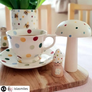 May include: A white teacup and saucer set with colourful polka dots, a wooden mushroom, and a wooden gnome figurine with a teacup design. A matching polka dot vase with bee designs is in the background. The items are on a wooden tray.