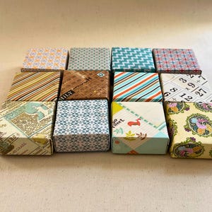 May include: A collection of small, square boxes with various patterned designs. The boxes feature geometric, floral, and abstract patterns in colors like blue, orange, and green. Some boxes have text. The boxes are arranged on a light-colored surface.