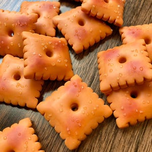 May include: Close-up of several orange, square-shaped crackers with a hole in the center and a scalloped edge. The crackers have a reddish-orange hue and are sprinkled with small, white salt crystals.