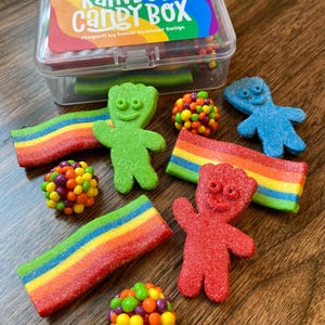 May include: A clear plastic box labeled "Rainbow Candy Box" with a variety of colorful candies. The candies include rainbow-colored sour belts, gummy bears, and small, round candies in various colors. The box is on a wooden surface.