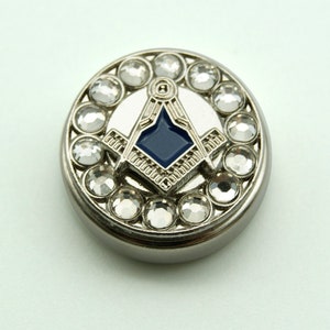 May include: A silver-coloured round button featuring a Masonic emblem. The emblem has a square and compass design with a blue centre, encircled by clear crystals. This button is likely for decorative use.