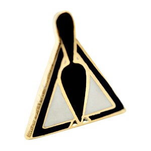 Può includere: Una spilla dorata con un design geometrico. La spilla presenta una combinazione di colori nero, bianco e oro. La forma è un triangolo con una forma a goccia nera al centro.