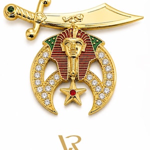May include: A gold-coloured pin featuring a sword, a crescent moon shape with embedded clear crystals, and a star with a red centre. The pin also includes a detailed depiction of an Egyptian pharaoh's head with red and green accents.