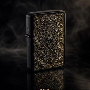 Könnte beinhalten: Ein mattschwarzes, rechteckiges Feuerzeug mit detaillierten, goldfarbenen Filigran-Gravuren auf der Vorderseite. Das Design zeigt ein zentrales Winkel- und Zirkelsymbol mit dem Buchstaben G. Der Schriftzug Victoria Regalia steht am unteren Rand.