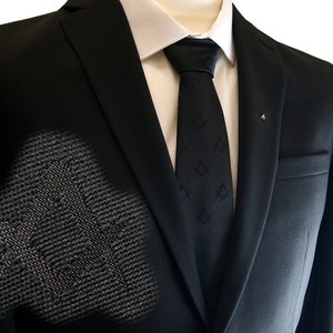 Op de afbeelding: Een zwart colbert met een zwarte stropdas met een herhalend patroon. Het colbert heeft een enkele knoopsluiting en een zak aan de rechterkant.