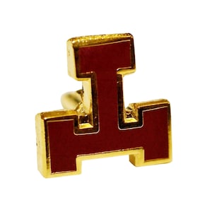 Op de afbeelding: Een gouden speld met een rood emaille ontwerp. Het ontwerp is een gestileerde letter "I" met een vierkante vorm boven en onder.