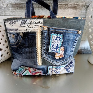 May include: A denim tote bag with a patchwork design. The bag features a dark blue denim exterior with a pocket, lace trim, and fabric accents. The handles are made of dark blue denim. The text "Westchester" is visible on the front.