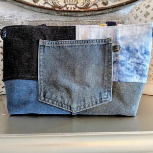 May include: A patchwork denim tote bag with a front pocket. The bag features panels of dark blue, medium blue, and light blue denim. The bag has a rectangular shape and a sturdy construction, perfect for carrying various items.