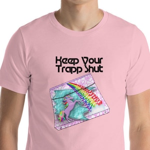 May include: A pale pink t-shirt with the words "Keep Your Trapp Shut" in black. The shirt has a graphic of a unicorn with a rainbow mane and a rainbow-coloured object. The unicorn is inside a box with a rainbow-coloured object.