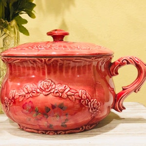 May include: A pink ceramic jar with a lid, featuring a floral design with raised roses and leaves. The jar has a handle and a decorative rim.