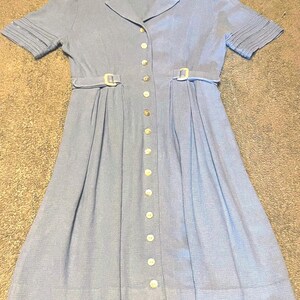 May include: A light blue button-down dress featuring short sleeves with pleated cuffs, a collared neckline, and a waistline accented by two light-colored rectangular buckles. The dress has a full-length front button closure with round buttons.