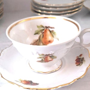 May include: A white teacup and saucer with a gold rim. The teacup and saucer feature a pear design with brown and green leaves.