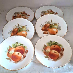 May include: Set of six white porcelain dessert plates with gold trim. Each plate features a different fruit design, including pears, apples, and grapes.