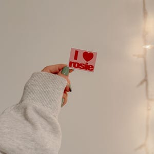 May include: A pink square sticker with the text "I ❤️ rosie" in red. The sticker is held up against a white wall. The text is in a bold, sans-serif font. The heart symbol is red.
