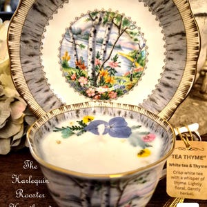 May include: A teacup and saucer set with a birch tree design, filled with a white liquid and decorated with flowers. The saucer and cup have gold trim. A tag reads "TEA THYME" with a description of the tea. The text "The Harlequin Rooster Hearth and Door" is also visible.